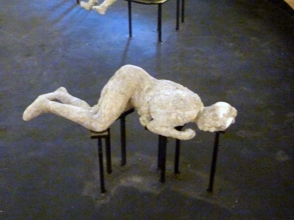 Victim 54. Cast of a victim made between 31st of July and 21st of September 1976.  
Plaster cast of victim on exhibit in display in temporary pyramid in amphitheatre. September 2015.
Victim 54 is a female aged over 20 years.
The body was found outside Porta Nola, by the Tomb of M. Obellius Firmus, between the 31st of July and the 21st of September 1976.
The imprint of the clothes, perhaps a tunic and a cloak, rolled up between the thighs, on the abdomen and back is clearly visible; on the right shoulder the drapery is evident and on the left calf the imprint of the weave of a light fabric seems to be distinguishable; there is possibly the presence of a headdress and sandals, traces of which appear to be visible on the left foot.
On the 15th of September 1976, next to the left hand of the victim part of an iron ring with a gem incised with an image of the goddess Fortuna.
On the 31st July, before the cast was made, near the body were found a gold ring with an emerald, a silver ring with the head of a serpent, another gold ring with bezel bearing the engraving of a palmette or ear of corn, a small silver ring with a chain to which a phallic pendant must have been hung, a silver statuette of Isis Fortuna and a hoard of money consisting of four silver denarii and twenty bronze coins.
See Osanna, N., Capurso, A., e Masseroli, S. M., 2021. I Calchi di Pompei da Giuseppe Fiorelli ad oggi: Studi e Ricerche del PAP 46, p. 446-448, Calco n. 54.

