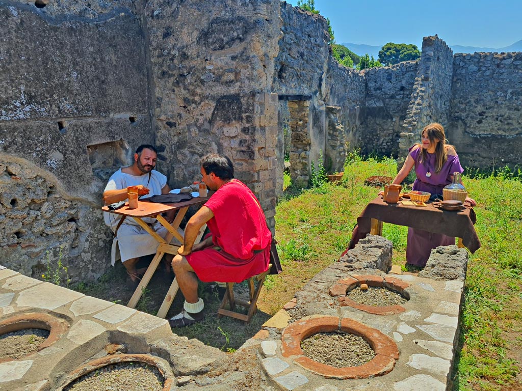 I.2.18 Pompeii. 8th June 2024. Looking across counter towards east wall and rear of bar at I.2.19. Photo courtesy of Giuseppe Ciaramella.
Historical reconstruction entitled L’altra Pompei prende vita (The other Pompeii comes to life).
