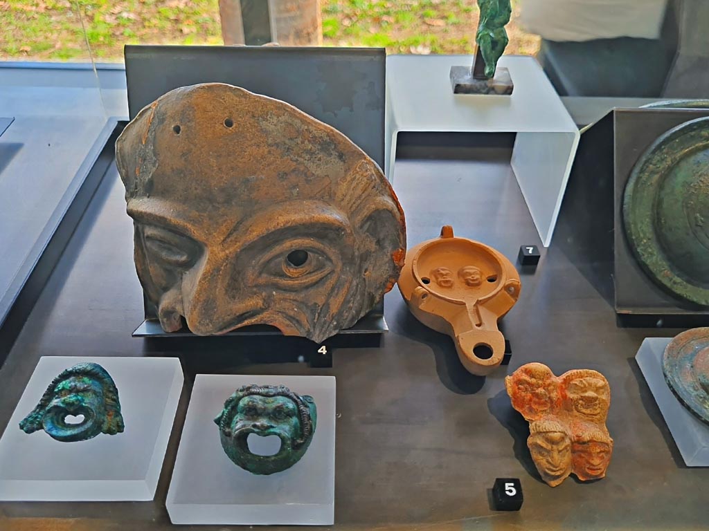 II.7.9 Pompeii. Palaestra. 
On display in exhibition in Palaestra entitled – “L’altra Pompei, vite comuni all’ombra del Vesuvio”. March 2024.
Photo courtesy of Giuseppe Ciaramella.
No.3, lower left – Bronze wall sconces with theatre masks, provenance not known, but from Pompeii. (Ist century AD).
No.4 – Terracotta theatre masks Pompeii – provenance not known, but from Pompeii. (Ist century AD). 
No.5 – Plaque with theatre masks, (Ist century BC – Ist century AD), from I.9.13.
No.6 (number hidden, but beneath No.7) – Oil lamp with theatre masks, from I.9.9.
On display in exhibition in Palaestra entitled – “L’altra Pompei, vite comuni all’ombra del Vesuvio”.

