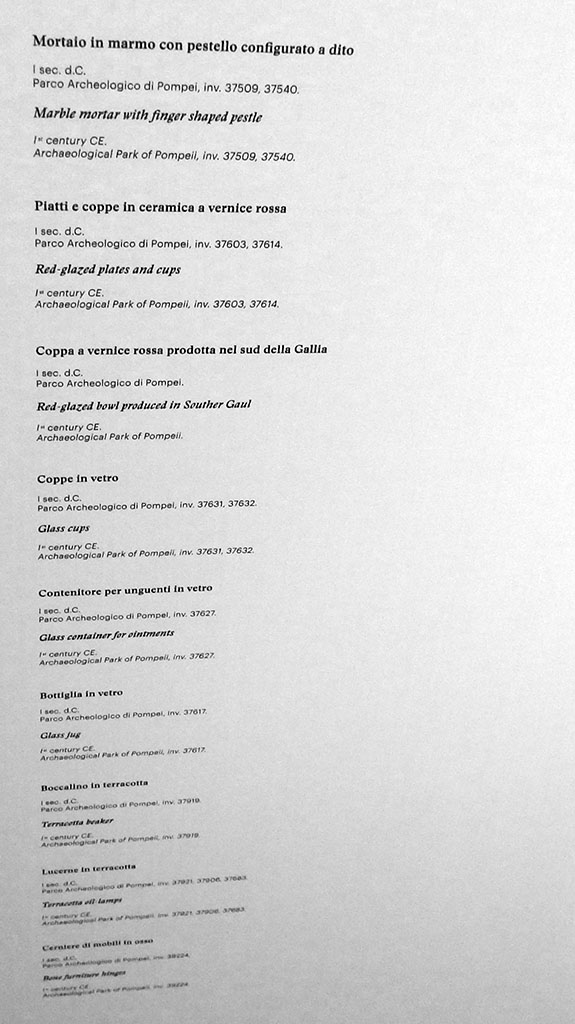 II.9.5 Pompeii, May 2018. Display notice on cabinet. Photo courtesy of Buzz Ferebee.
Marble mortar with finger shaped pestle (Mortaio in marmo con pestello configurato a dito), inv. nos. 37509, 37540
Red-glazed plates and cups (Piatti e coppe in ceramica a vernice rossa), inv. nos. 37603, 37614
Red-glazed bowl produced in Southern Gaul (Coppa a vernice rossa prodotta nel sud della Gallia),
Glass cups (Coppe in vetro), inv. nos. 37631, 37632
Glass container for ointments (Contenitore per unguenti in vetro), inv. no. 37627
Glass jug (Bottiglia in vetro), inv. no. 37617
Terracotta beaker (Boccalino in terracotta), inv. no. 37919
Terracotta oil lamps (Lucerne in terracotta), inv. nos. 37921, 37906, 37683
Bone furniture hinges (Cerniere di mobili in osso), inv. no. 39224
All inventory numbers are from Archaeological Park of Pompeii.
