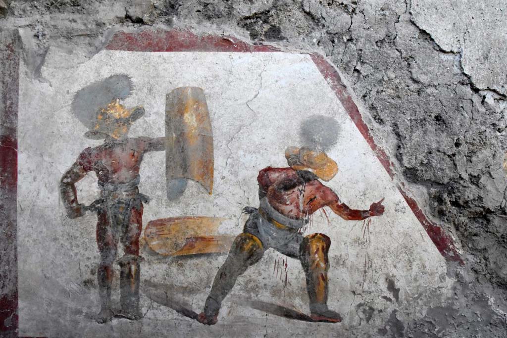 A contest between a Mirmillo and a "Thracian", two types of fighters distinct from different armour and classic opponents in gladiatorial fights. 
On a white background, bordered on three sides by a red band, the scene of struggle between the two gladiators is composed. 
On the left is a "Mirmillo" of the "Scutati" category: he wields the offensive weapon, the gladium, (short Roman sword) and a large rectangular shield (scutum) and wears a wide-brimmed helmet equipped with visor with plumes, the cimian. 
The other, in a succumbing position, is a "Thracian", a gladiator of the "Parmularii" category, with the shield on the ground. 
He is represented with a helmet (galea), with wide brim and wide visor to protect the face, topped by a tall crest.

Si tratta della scena di un combattimento tra un “Mirmillone” e un “Trace”, due tipologie di lottatori distinti da armature differenti e classici avversari nelle lotte gladiatorie.
Su uno sfondo bianco, delimitato su tre lati da una fascia rossa, si compone la scena di lotta tra i due gladiatori. Quello sulla sinistra è un “Mirmillone” della categoria degli "Scutati”: impugna l’arma di offesa, il gladium, (corta spada romana) e un grande scudo rettangolare (scutum) e veste un elmo a tesa larga dotato di visiera con pennacchi, il cimiero. L’altro, in posizione soccombente, è un “Trace”, gladiatore della categoria dei "Parmularii”, con lo scudo a terra. È rappresentato con elmo (galea), a tesa larga ed ampia visiera a protezione del volto, sormontato da un alto cimiero.

Photograph © Parco Archeologico di Pompei.

