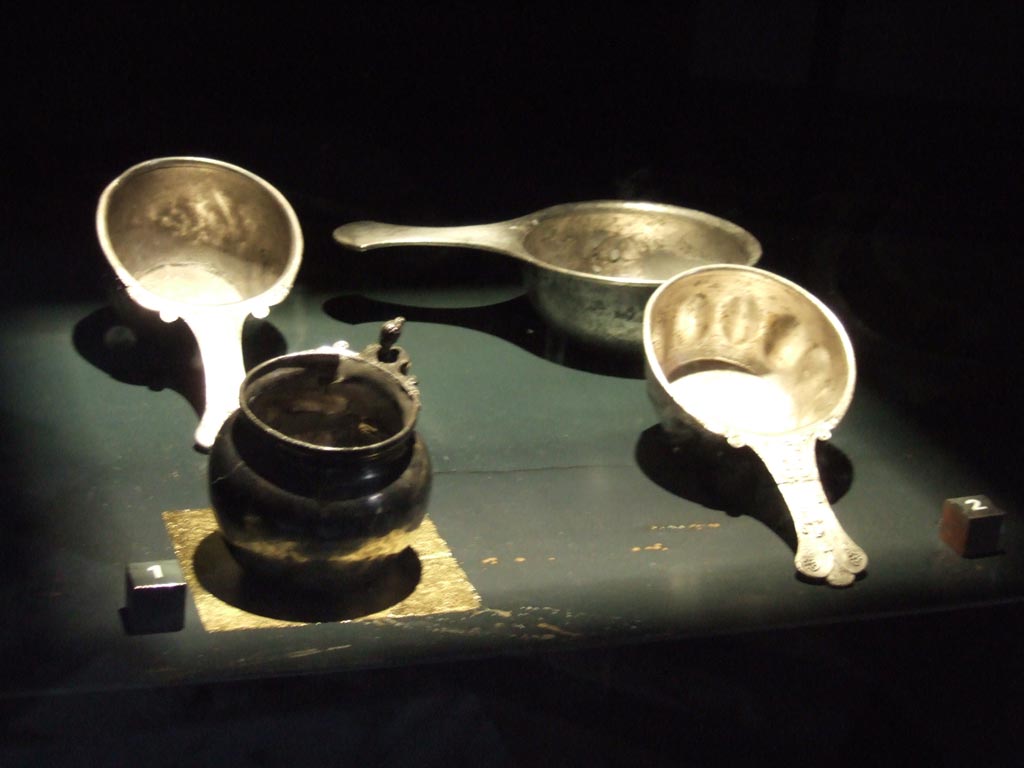 VI.12.2 Pompeii. Silver found in VI.12.2. Now in Naples Archaeological Museum.
Urceolus or small jug or pitcher (left front) with female head. Inventory number 25680.
Patera or drinking vessel (right front) with indented pattern on the sides of the bowl and naked figure of Mercury on handle. Inventory number 25338.
Patera (Left rear) with the handle decorated with the head of a faun. Inventory number 25340.
Patera (Right rear) with less ornate handle. Inventory number 25339.
See Guzzo, P. (A cura di), 2006. Argenti a Pompei. Milano, Electa. (p. 112-113, Nos. 102-5).
