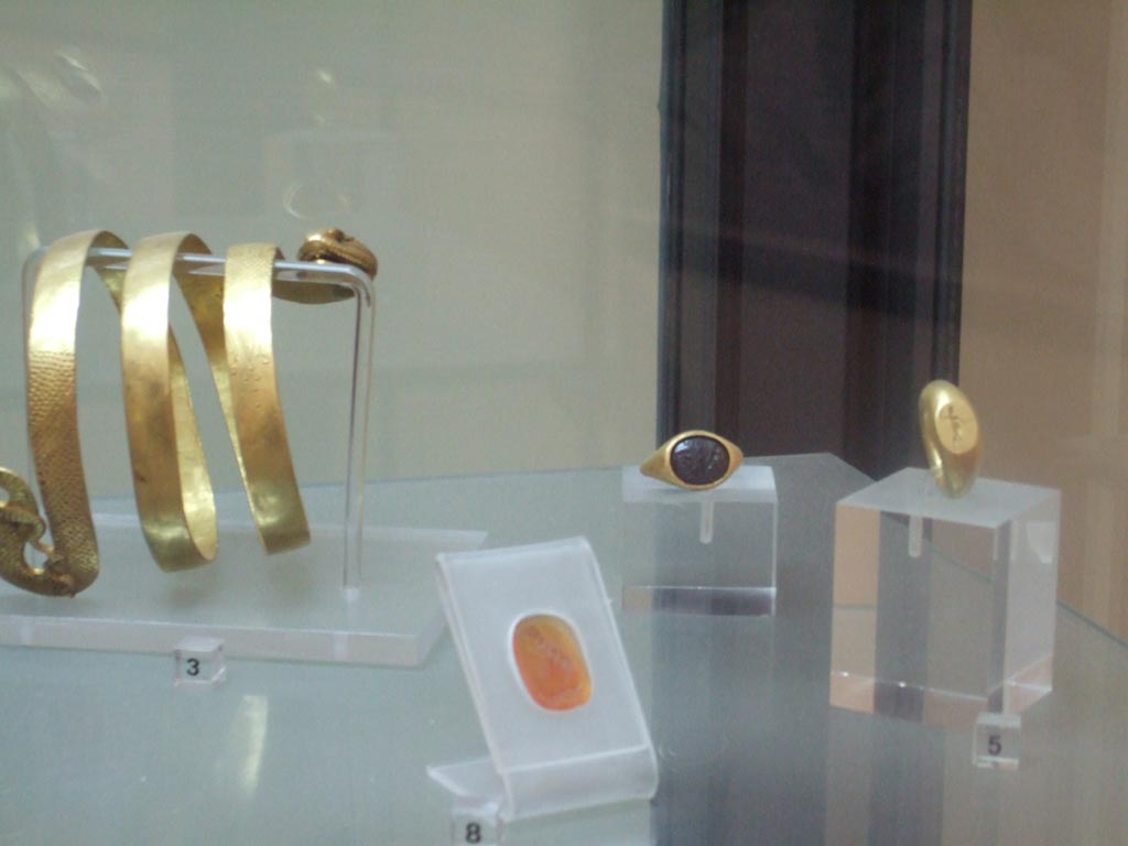 VI.12.2 Pompeii. Found 4th March 1831. Jewellery found in VI.12.2. 
Carving on cornelian of bust of a youth, possible Alexander the Great (in front). Inventory number 26766.
Gold ring with oval centre representing Hercules in the garden of Hesperides (on right). Inventory number 25132.
Gold ring with nude figure, with discus/shield and javelin (at rear). Inventory number 25084.
Now in Naples Archaeological Museum. 
