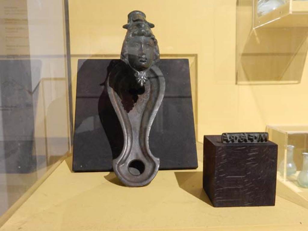 VI.17.42, Pompeii, May 2018. Oil lamp in bronze with feminine face on the bottom part of the handle.
Parco Archeologico di Pompei, inventory number 13373.
On the right is the seal of Marcus Fabius Rufus, which according to the information card was found in this house.
Photo courtesy of Buzz Ferebee.