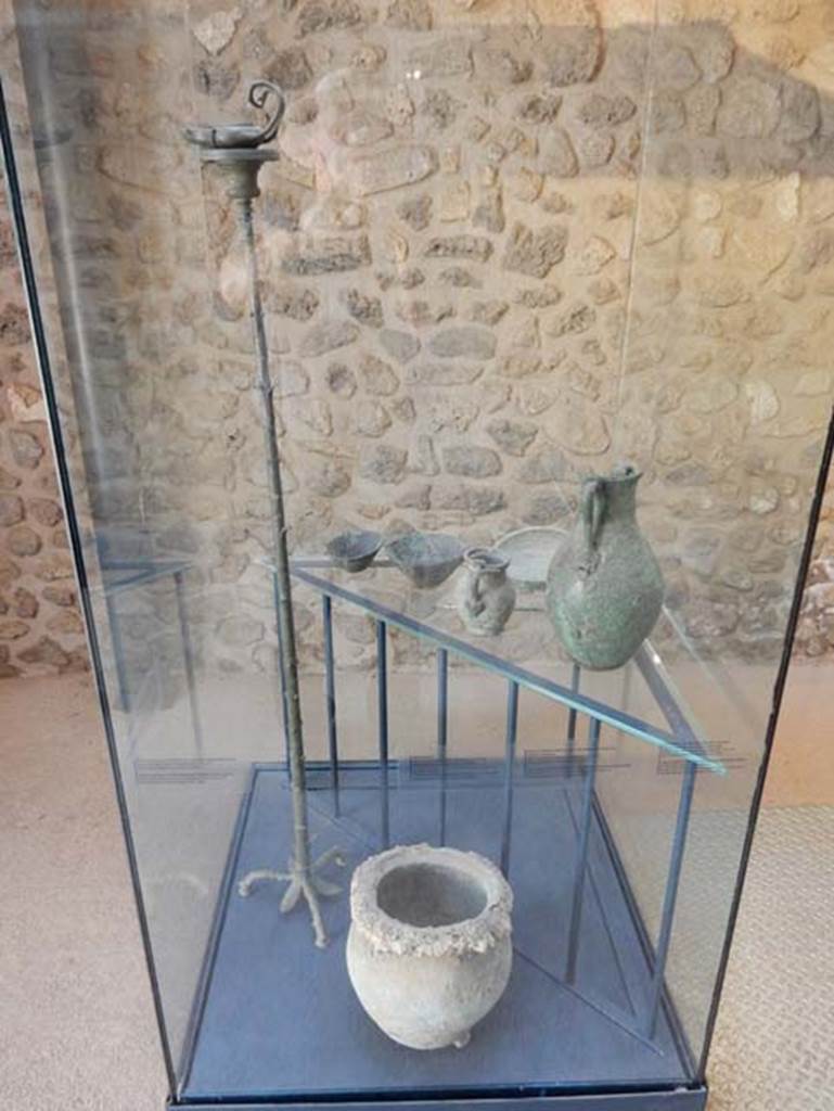 VIII.2.1 Pompeii. May 2018. Display case with finds from excavations conducted in the area since the 1930’s.
The top shelf shows a bronze needle, a bronze jug with female face, a small bronze amphora decorated with theatre masks and three bronze cups.
Bottom is a bronze olla (pot used for boiling), which conserves some of the lapilli from the eruption. Pompeii inventory 6322.
Also in the case is a bronze lamp on a tall stand.
All were found in the area to the south of the House of Championnet.
Photo courtesy of Buzz Ferebee.

