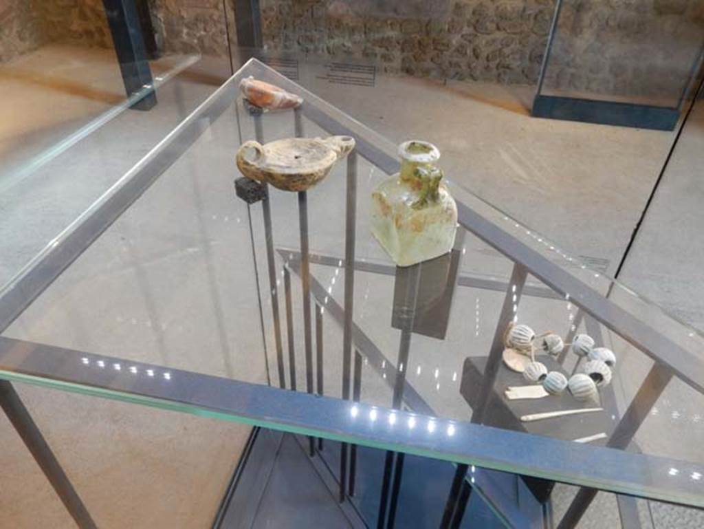 On display in Championnet complex  Pompeii. May 2018. Finds from excavations conducted in the area since the 1930’s.
Elements of a glass paste necklace. Pompeii inventory number 6330.
Green glass bottle. Pompeii inventory number 14828.
Two terracotta lamps.
Found in the area south of the House of Championnet.
Photo courtesy of Buzz Ferebee.
