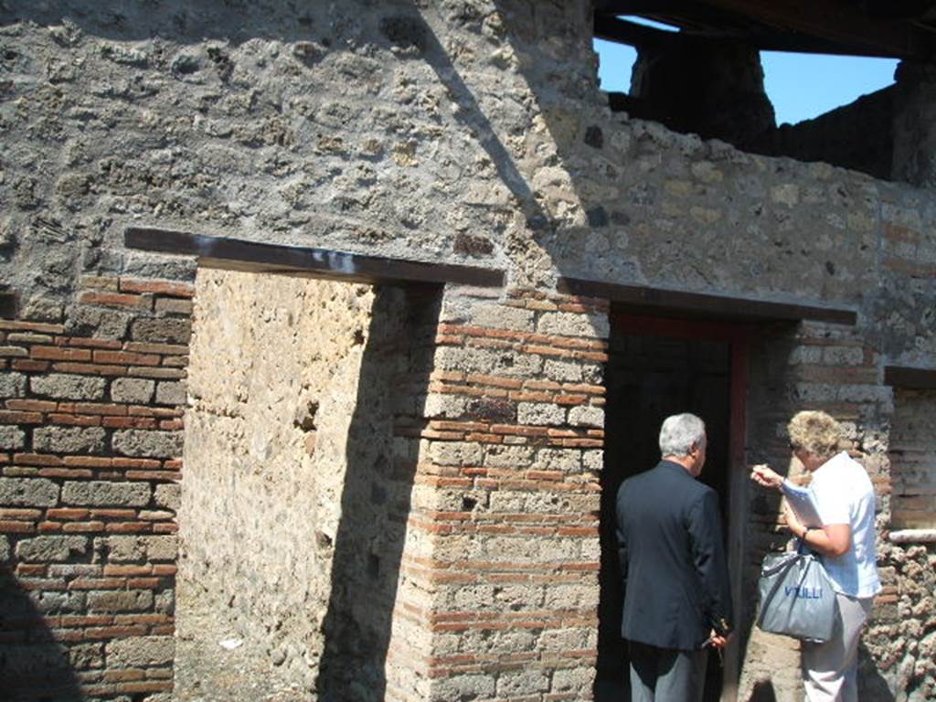 IX.5.16 Pompeii. May 2005. West side of atrium a’, on the left, doorway to kitchen and latrine e’.
On the right, doorway to cubiculum f’.
According to Boyce, on the south wall of the kitchen was a large rectangular panel of white stucco.
On it were painted two yellow serpents with red crest and beards. 
These were on either side of a tile embedded and projecting from the wall.
A fire was painted on the wall above the tile.
The background was painted with plants, and across the top were two painted garlands.
On each of the garlands was a painted bird.
See Boyce, G. K., 1937. Corpus of the Lararia of Pompeii. Rome: MAAR 14.  (p. 86, no. 425)

In IX.5.16, the first place on the left of the atrium was searched again, and found were several iron nails, some fish bones, and in terracotta: two containers for oil, a vase in fragments, two amphorae, two pot lids, and a broken bowl containing ash.
See Notizie degli Scavi di Antichità, 1885, (pp. 162-4).

