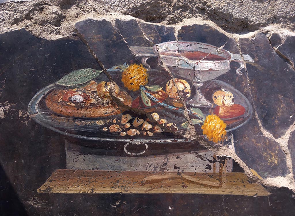 IX.10.1 Pompeii. June 2023. Atrium 2 west wall. Still life.
The composition, created in a free field in the centre of a panel with a black background enclosed by a carpet border, is structured on two perspective planes rendered by shelves. On the upper shelf rests a large silver tray with movable handles containing a cantharus of the same material, filled with wine, and various dried and seasonal fruits. Part of these fruits are painted above a brown element (perhaps a basket or, more likely, a focaccia) which occupies the entire left portion of the tray. The lower shelf rendered in perspective evokes a wooden canteen on which hangs a band hanging from the fruits of the composition.
IX.10.1 Pompeii. Giugno 2023. Atrio 2 parete ovest. Natura morta.
La composizione, realizzata in campo libero al centro di un pannello a fondo nero racchiuso da una bordura di tappeto, è strutturata su due piani prospettici resi da mensole. Sulla mensola superiore è poggiato un grande vassoio in argento ad anse mobili contenente un cantharus dello stesso materiale, ricolmo di vino, e diversi frutti secchi e di stagione. Parte di questi frutti sono dipinti al di sopra di un elemento di colore bruno (forse una cesta o, più probabilmente, una focaccia) che occupa l’intera porzione sinistra del vassoio. La mensola inferiore resa in prospettiva rievoca una mensa in legno su cui ricade una benda pendente dai frutti della composizione.
Photograph © Parco Archeologico di Pompei.
Vedi/See Una natura morta con xenia dallo scavo della casa IX 10,1 a Pompei. E-journal 3, Scavi di Pompeii, 27.6.2023, p. 2, fig. 2. E-journal on PAP web site

