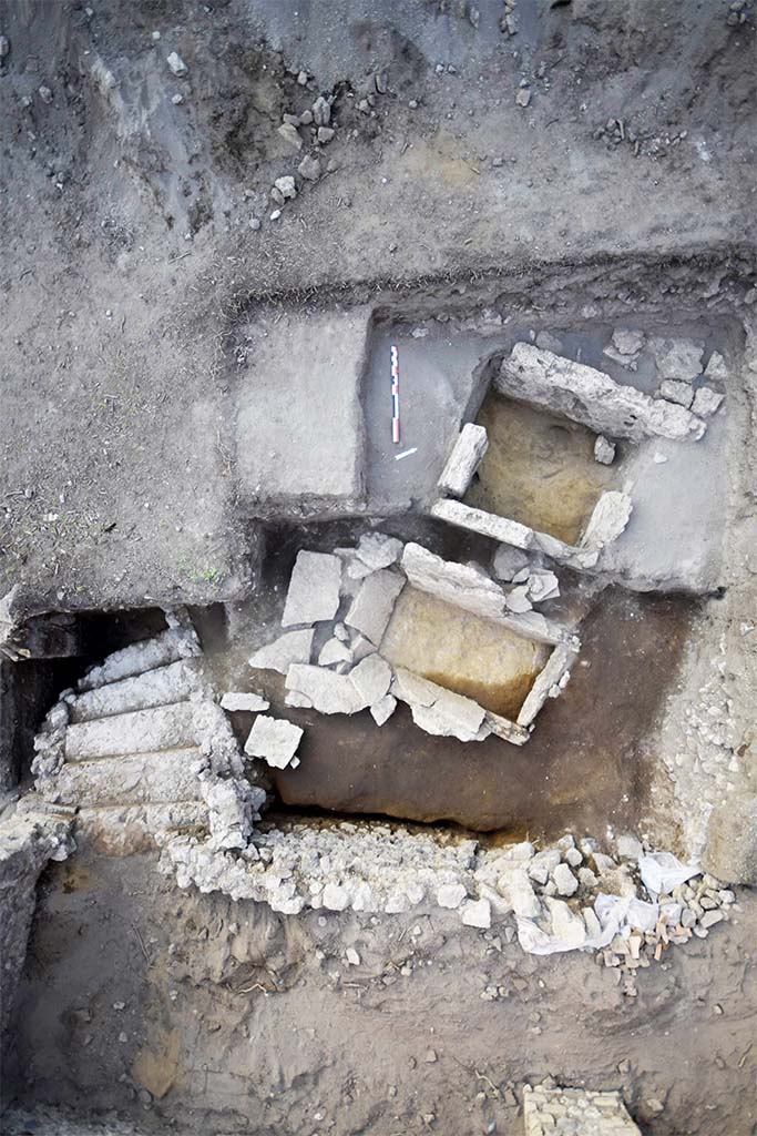 HGE31B with one side missing in 2015. HGE31A is below and the still to be uncovered HGE32A above.
HGE30 is at the bottom with the stairs of the shop at the bottom left.
Photo � B. Lemaire, EFR/CNRS.

Separated by 0.60 m from the previous one and nearly 0.50 m from the third, this grave is completely filled by reworked fill (Figs. 15 and 23). Only three Sarno limestone slabs are preserved and although the north-western one is missing, the space thus delimited is 1.44 m from north-east to south-west and nearly 0.75 m restored from north-west to south-east. The slabs measure between 0.92 and 0.99 m in height (up to 32.30 m s.l.m.) and only the one in the north-east measures only 0.62 m, but is set on wedge stones of about 10 cm. (30102). 

This burial structure was excavated in the modern period, as evidenced by the levels of fill found inside. It is only a single fill. Finally, the space observed was over-excavated by our predecessors, deeper than the installation levels, making it possible to observe the foundation of the slabs and even the wedging elements of one of them. No original floor and fill levels could be observed, nor any elements relating to the remains of the deceased or to offerings.

See CEFR 1581 or para 46 and 47 below
