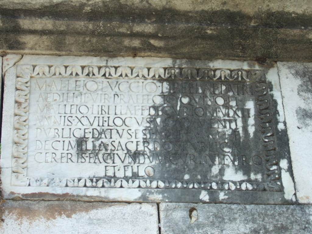 HGE37 Pompeii. May 2006. Marble plaque on east side. On the plaque is the inscription:
M. ALLEIO LVCCIO LIBELLAE PATRI 
AEDILI II VIR PRAEFECTO QVINQ(uennali) ET 
M. ALLEIO LIBELLAE F DECVRIONI VIXIT 
ANNIS XVII LOCVS MONVMENTI
PVBLICE DATVS EST ALLEIA M F 
DECIMILLA SACERDOS PVBLICA 
CERERIS FACIVNDVM CVRAVIT VIRO 
ET FILO

This differs from the south side plaque where the last word is FILIO rather than FILO.
The plaque is also narrower and the inscription is on more (eight) lines.
According to Epigraphik-Datenbank Clauss/Slaby (See www.manfredclauss.de) this reads

M(arco) Alleio Luccio Libellae patri
aedili IIvir(o) praefecto quinq(uennali) et 
M(arco) Alleio Libellae f(ilio) decurioni vixit 
annis XVII locus monumenti
publice datus est Alleia M(arci) f(ilia)
Decimilla sacerdos publica
Cereris faciundum curavit viro
et filio      [CIL X 1036 second part]

Gell describes this as “A well-executed tomb, in stone, of simple form, about 15 feet high. 
Upon two of its sides are similar inscriptions, which inform us that it was erected by Alicia Decimilla, public priestess of Ceres, to her husband Luccius Libella, aedile, duumvir, and quinquennial prefect : also to her son M. Alleius Libella, decurion at 17, upon ground decreed by the public for that purpose.”
See Gell, W, and Gandy J. P., 1819.  Pompeiana. London: Rodwell and Martin.  (p. 111).