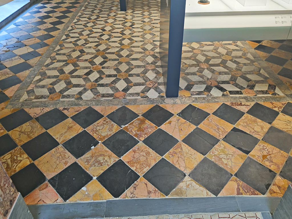 Mosaic in Naples Archaeological Museum, room CXXXVIII (138). June 2024. Photo courtesy of Giuseppe Ciaramella.
According to Miele –
“In the centre of the floor is a floor detached from the Villa Imperiale on Capri, surrounding it are the larger alternating slate and antique yellow squares, detached from an apsidal building next to the Second Complex.”

According to La Vega –
28 Ottobre 1775.
In Stabia si e quasi del tutto scavata la stanza notata in piu antecedenti rapporti; e questa rettangola e termina da un lato con semicircolo che l’abbraccia da due lati; la parte rettangola e di palmi 29 ed once 5 per palmi 31 ed once 5, ed il semicircolo ha di diametro palmi 48 ¼; le mura che la chiudono erano bene dipinte con fondo rosso, ma ora ne restano in piedi per solo otto o nove palmi d’altezza; il pavimento e di marmo con alternativi quadrati, uno di giallo antico ed altro di lavagnone, ed attorno al margine vi sono delle fasce di pietre di vari colori.
Li quadrati che fanno il pavimento della parte rettangola della stanza sono di lato once 14 e quelli che fanno il pavimento della parte semicircolare sono once 10. Subito che questo pavimento sara intieramente scoperto avvisero Canart che lo mandi a levare…………………
(In Stabia, the room noted in several previous reports has been almost completely excavated: it is rectangular and ends on one side with a semicircle that embraces it on two sides; the rectangular part is 29 palms and 5 onces by 31 palms and 5 onces, and the semicircle has a diameter of 48 ¼ palmi; the walls that enclose it were well painted with a red background, but now they remain standing only 8 or 9 palmis high; the floor is made of marble with alternative squares, one of Antique Yellow and the other of Lavagnone, and around the margin there are bands of stones of various colours.
The squares that make the floor of the rectangular part of the room are 12 inches, and those that make the floor of the semicircular part are 8 inches. As soon as the floor will be completely uncovered, I will advise Canart to lift it…….)
See Ruggiero M., 1881. Degli scavi di Stabia dal 1749 al 1782, Naples, (p.232).
