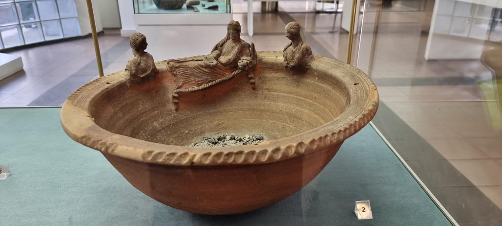 Boscoreale, December 2023. Villa rustica in via Casone Grotta, proprietà Risi di Prisco. Lararium.
On the left side of the niche, slightly more at the bottom, stuck in the wall, was a shelf.
On this was found an ordinary uncoloured clay ceramic basin.
The terracotta basin has three female figures on the rim. 
Two are busts and are facing the third, in the centre, who is lying on the rim.
Now in Boscoreale Antiquarium. Photo courtesy of Miriam Colomer.
It is significant that the motif of the female figure lying on the kline was repeated on the statuette, the silver sheet, as well as on the clay basin.
Regarding the latter, it should be noted that burnt material, so far unidentified, was found in it. 
It is assumed that the basin, placed on the altar during the cult ceremony, was later placed on the clay shelf next to the Lararium, where it was found. 
All the other objects found in the room were also likely to be used exclusively for domestic worship.
See Fergola, L., 1987. Comune di Boscoreale. Via Casone Grotta, proprietà Risi Di Prisco, in Rivista di Studi Pompeiani, 1, p. 164.
